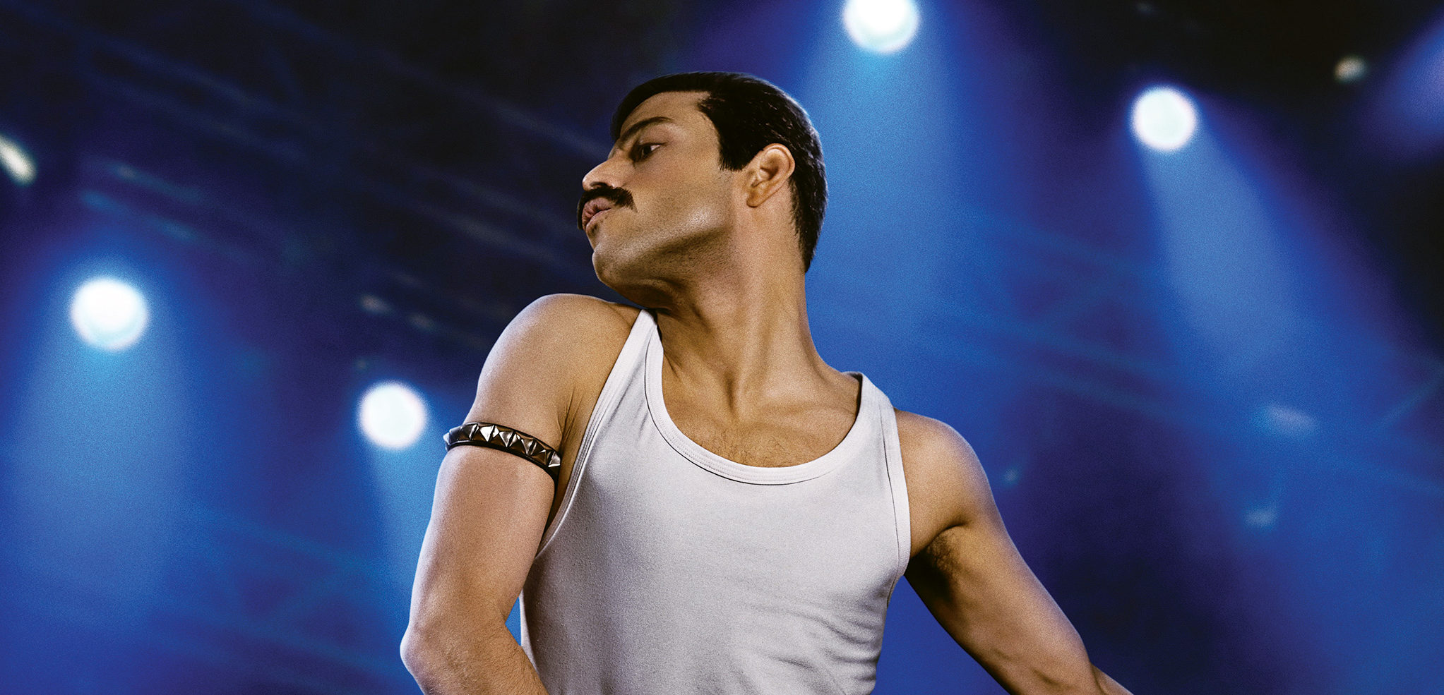 Rami Malek posed as Freddie Mercury in a white tank top and black, spiked armband. He's in front of a blue concert-lit background.