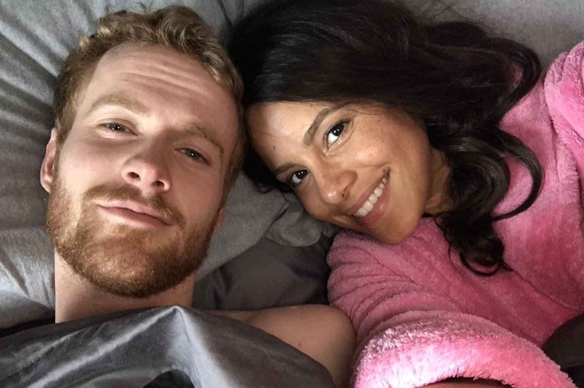 Parisa Fitz-Henley, right, and Murray Fraser take a selfie in costume as Prince Harry and Meghan Markle while lying down on a bed.