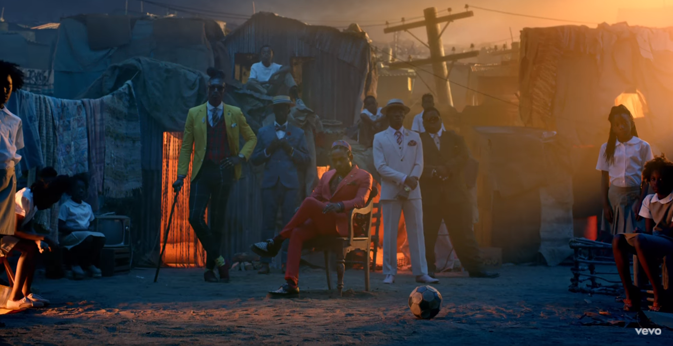 Kendrick Lamar sits in a red tailored suit among brightly-dressed men in a shanty town at sunset.