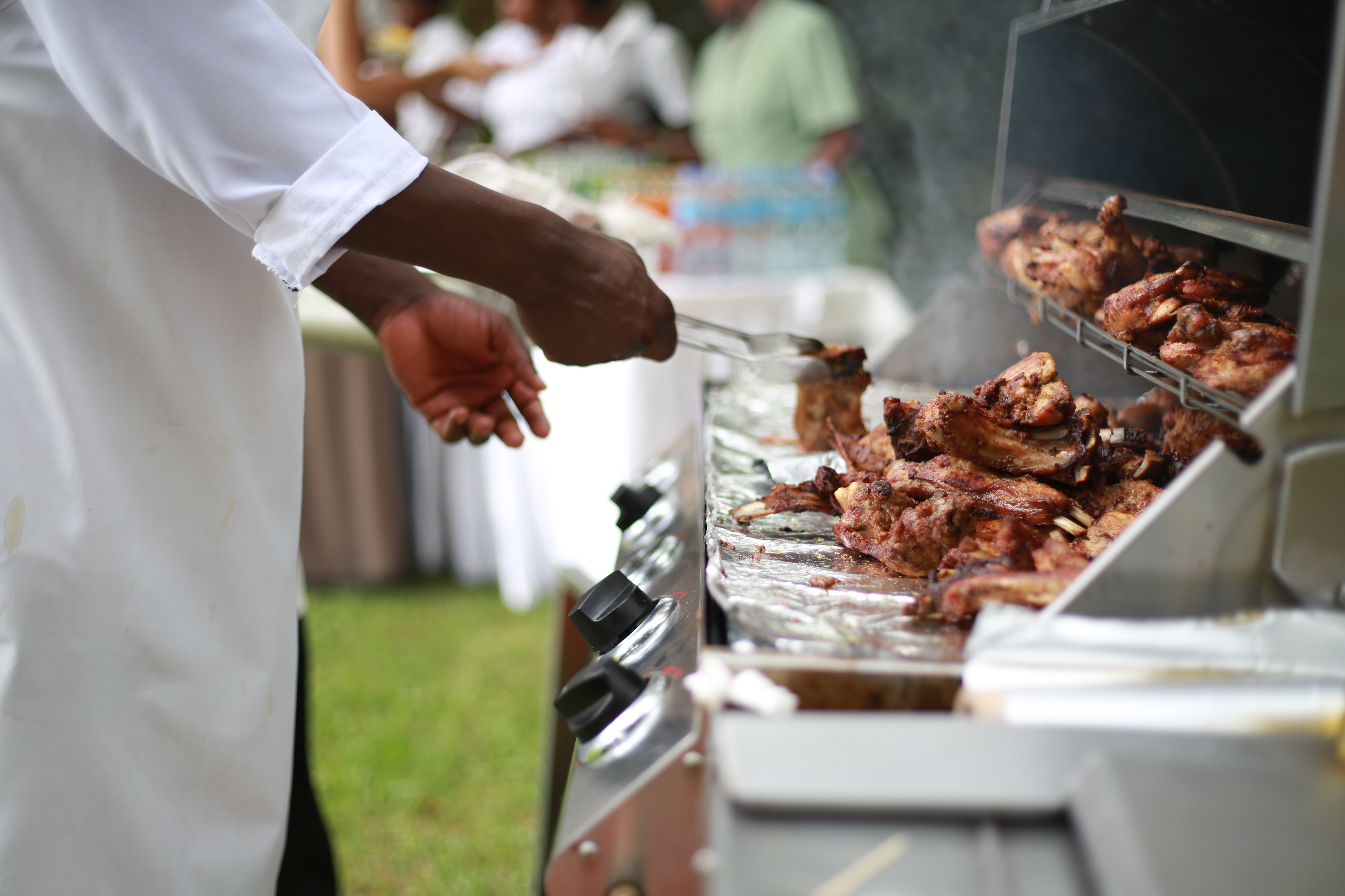 Image description: a black man flips grilled chicken on a stainless-steel grill on a summer day.