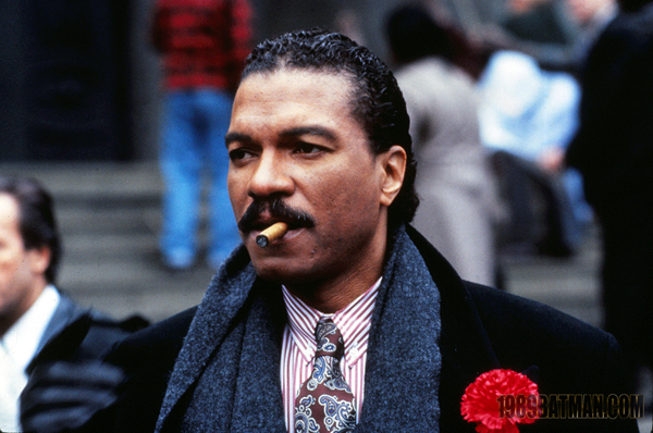 Billie Dee Williams as Harvey Dent is dressed in an expensive coat with red carnation as decoration. He's also wearing a grey scarf, purple and white pinstripe shirt, and purple and blue patterned tie. He has a cigar in his mouth. 