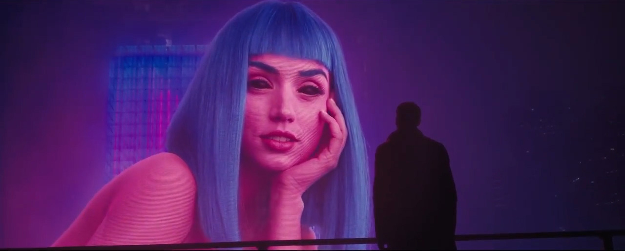 A huge holographic billboard of Joi, with pink-purple skin and blue hair, looks at K. 