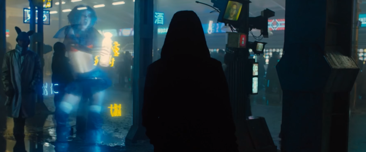 A harajuku girl dances in a hologram as a hooded figure walks by.
