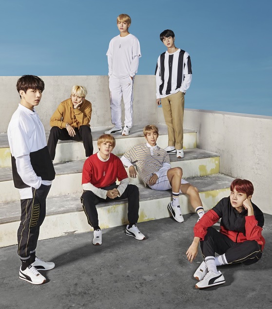 Members of BTS posed on white steps and partitions against a blue sky, wearing clothes from their line. 