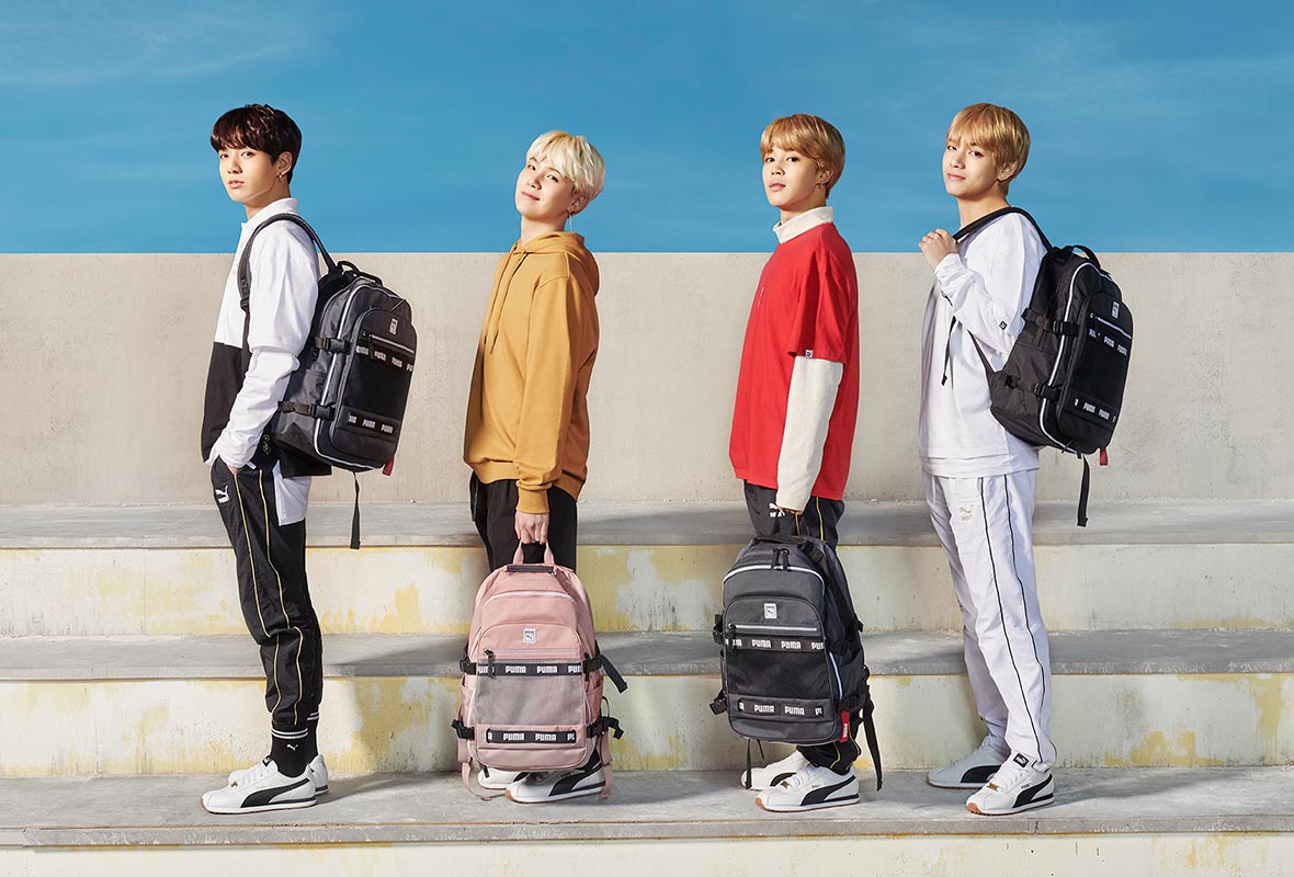 Members of BTS posed near white steps against a blue sky, carrying backpacks from their line. 