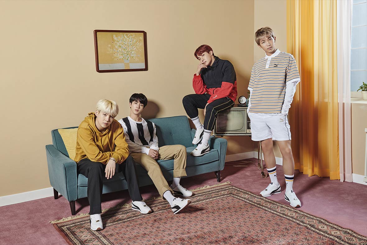 Members of BTS, wearing clothes from their line, posing in a set staged like a living room with a blue couch and faded burgundy rug. 