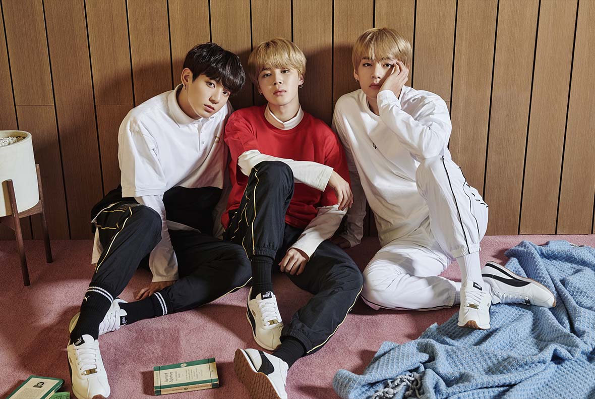 Members of BTS, wearing clothes from their line and sitting against a brown wood-paneled wall, posing in a set staged like a living room. 