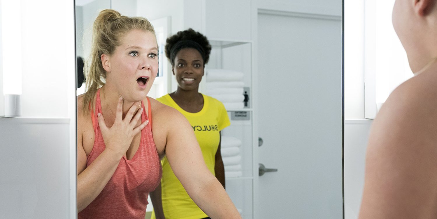 Amy Schumer reacts to her reflection in the gym bathroom mirror with Sacheer Zamata reacting to Schumer in the background in "I Feel Pretty"