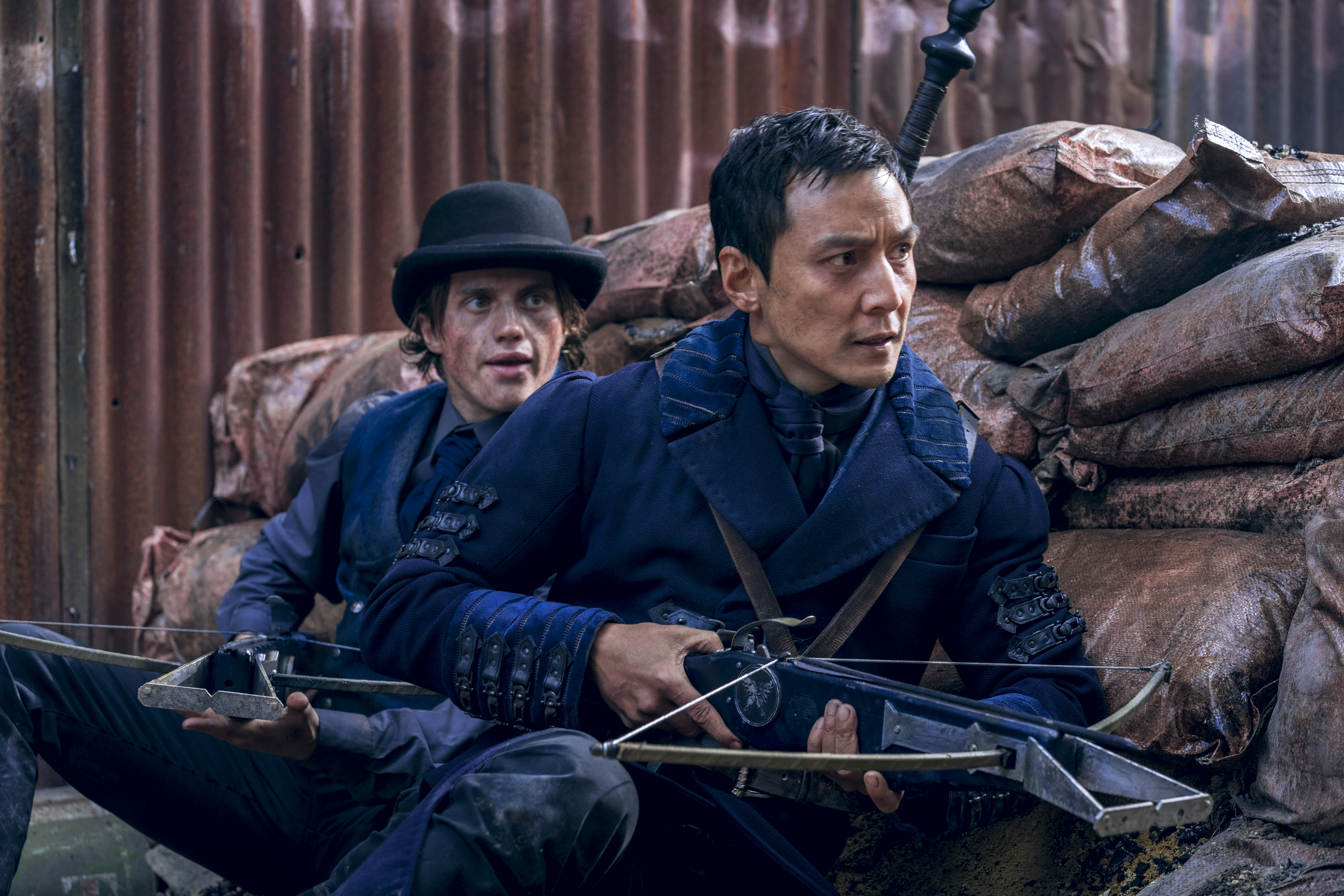 George Sear as Arthur, Daniel Wu as Sunny. They're crouched behind a makeshift barracks. Both are wearing blue velvet overcoats and Arthur is wearing a dark blue or black bowler hat. (Photo Credit: Aidan Monaghan/AMC)