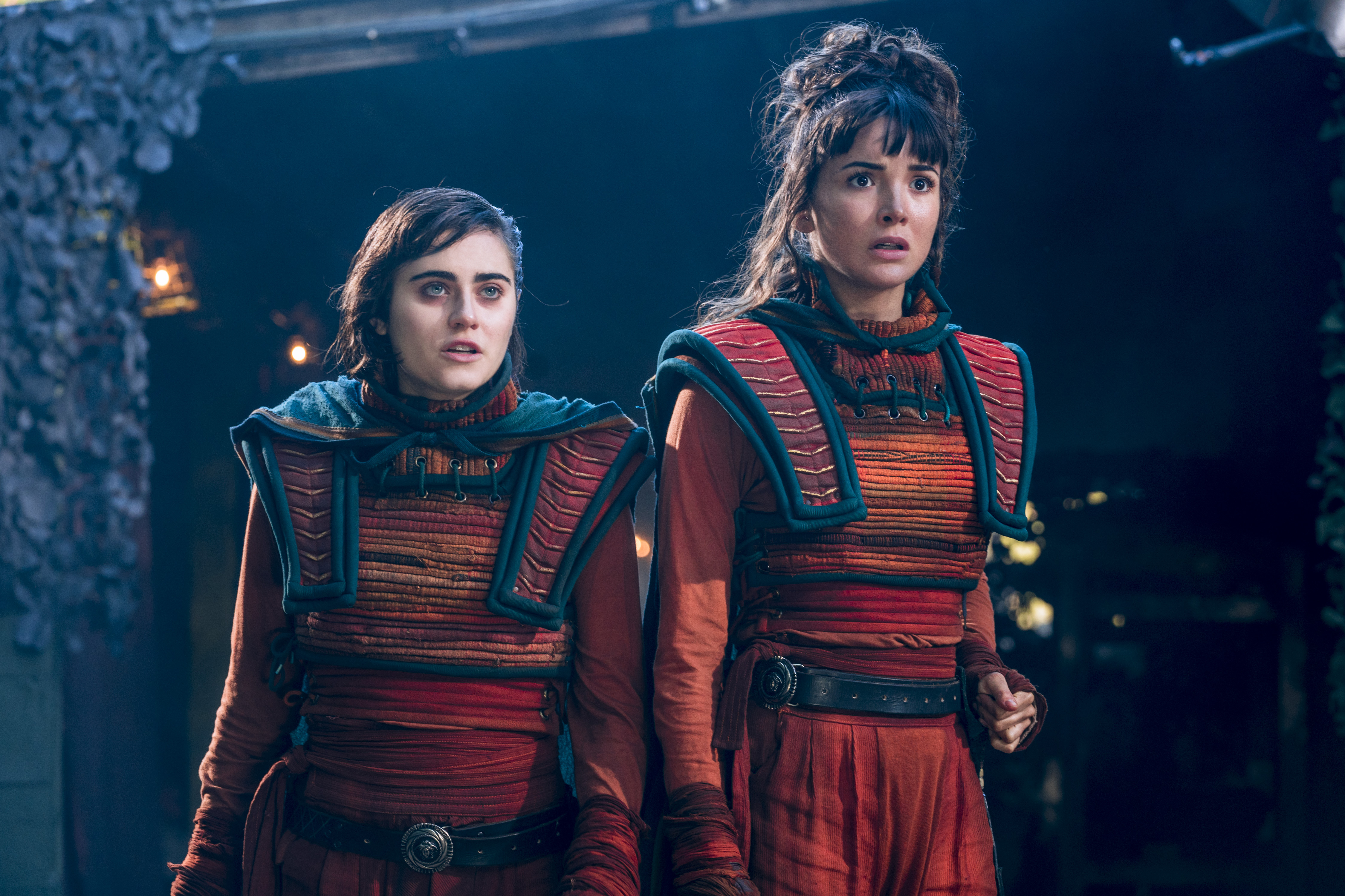 Ally Ioannides as Tilda, Maddison Jaizani as Odessa. Both are wearing orange woven outfits with red and blue accents. (Photo Credit: Aidan Monaghan/AMC)