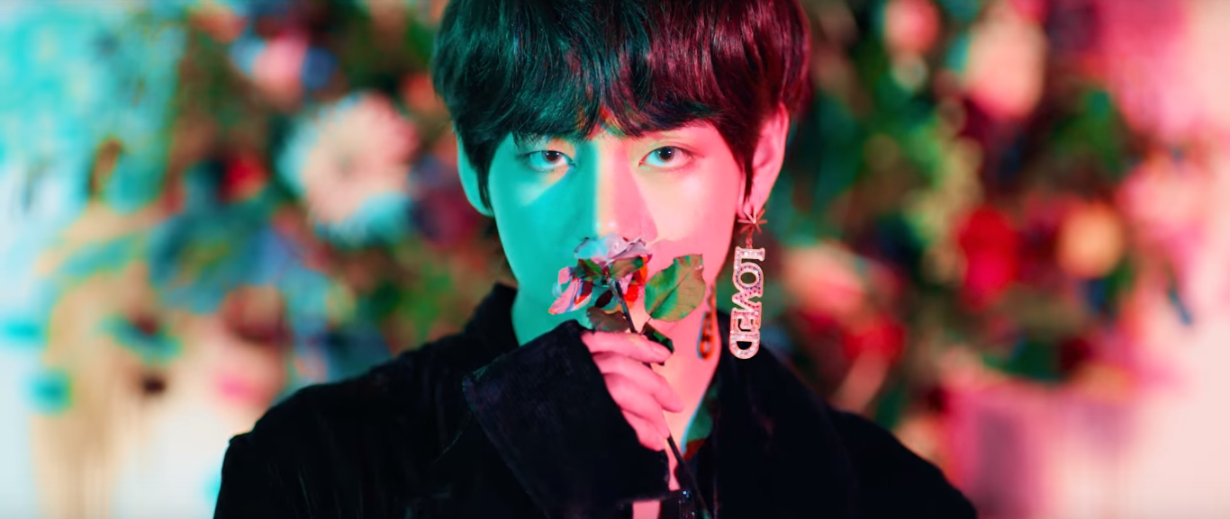 Taehyung lit by green and pink lights, holds a rose to his nose. Hes wearing a black velvet oversized jacket and wears an earring that spells "LOVED". He stands in front of a rosebush.