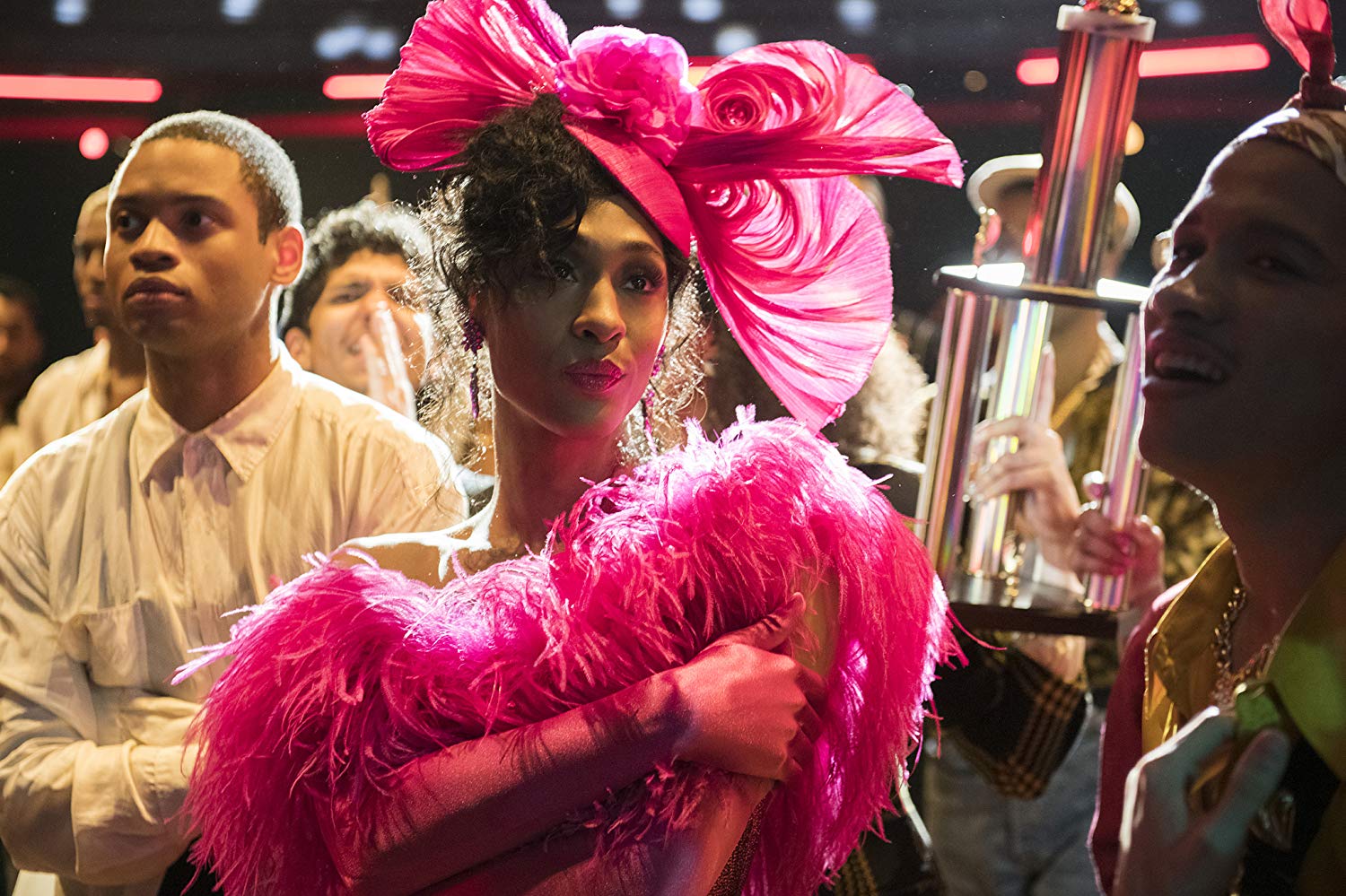 Amid a crowd, Mj Rodriguez, in a feathery pink outfit and wearing a pink hat, holds a trophy after winning a ball category.