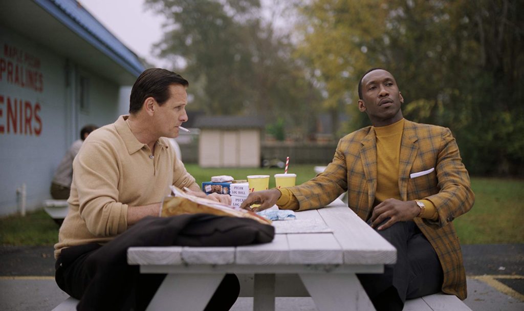Viggo Mortensen as Tony Lip and Mahershala as Dr. Shirley, sitting at a picnic table eating fast food. Dr. Shirley looks off into the distance as Tony Lip smokes a cigarette. 