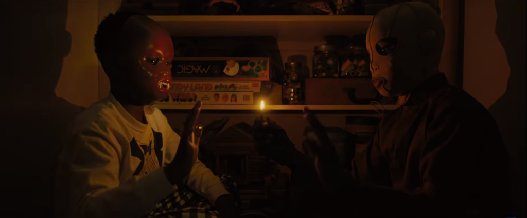 Evan Alex and his doppleganger sit in a flame-lit room. Evan wears a Chewbacca mask and his evil self wears a white mask. Both are holding a hand up to each other. 