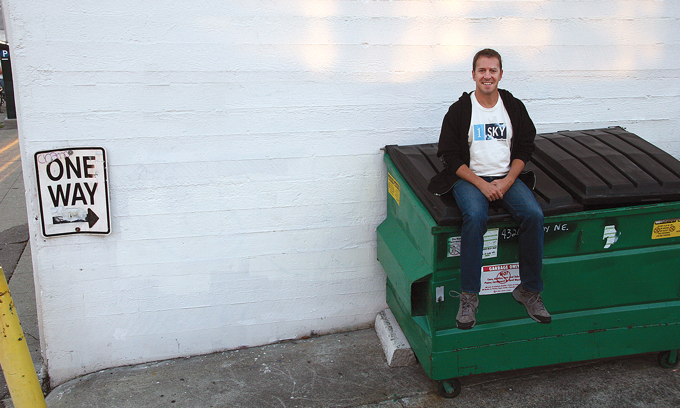 Colin Beavan sits on the top of a recycling bin outside in front of a whitewashed wall.