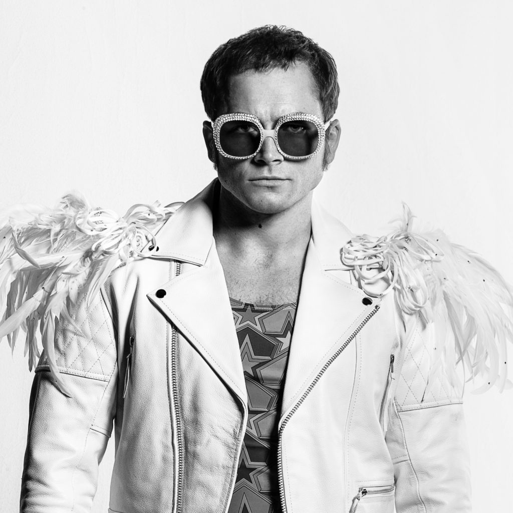 Taron Egerton as Elton John in Rocketman from Paramount Pictures. The black and white picture features Egerton in costume with a white leather jacket with white plumes on the shoulders and large bejeweled sunglasses. 