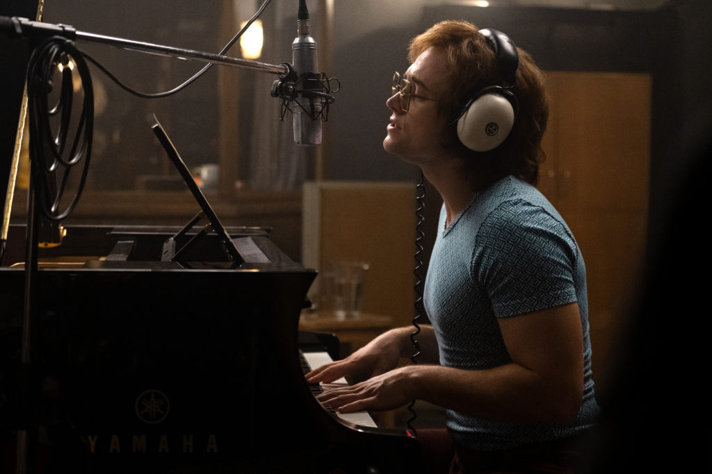 Taron Egerton as Elton John in Rocketman from Paramount Pictures. He's wearing a blue fitted t-shirt and headphones as he records himself playing the piano.