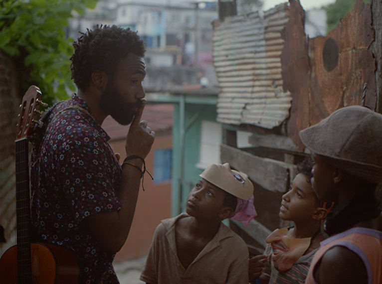 Donald Glover in Guava Island. Glover is jauntily telling some kids in masks to shush with his finger on his mouth. He has a guitar on his back and wears a blue shirt with blue and pink flowers. 