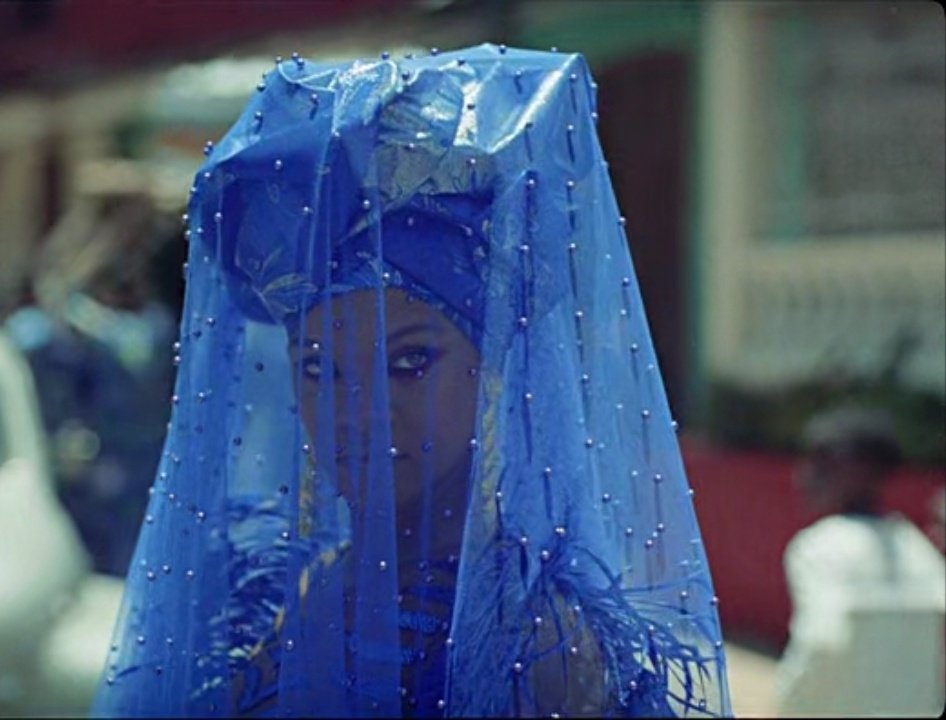 Rihanna as Deni's girlfriend Kofi. She wears a blue ornate dress with a blue turban and beaded veil. 