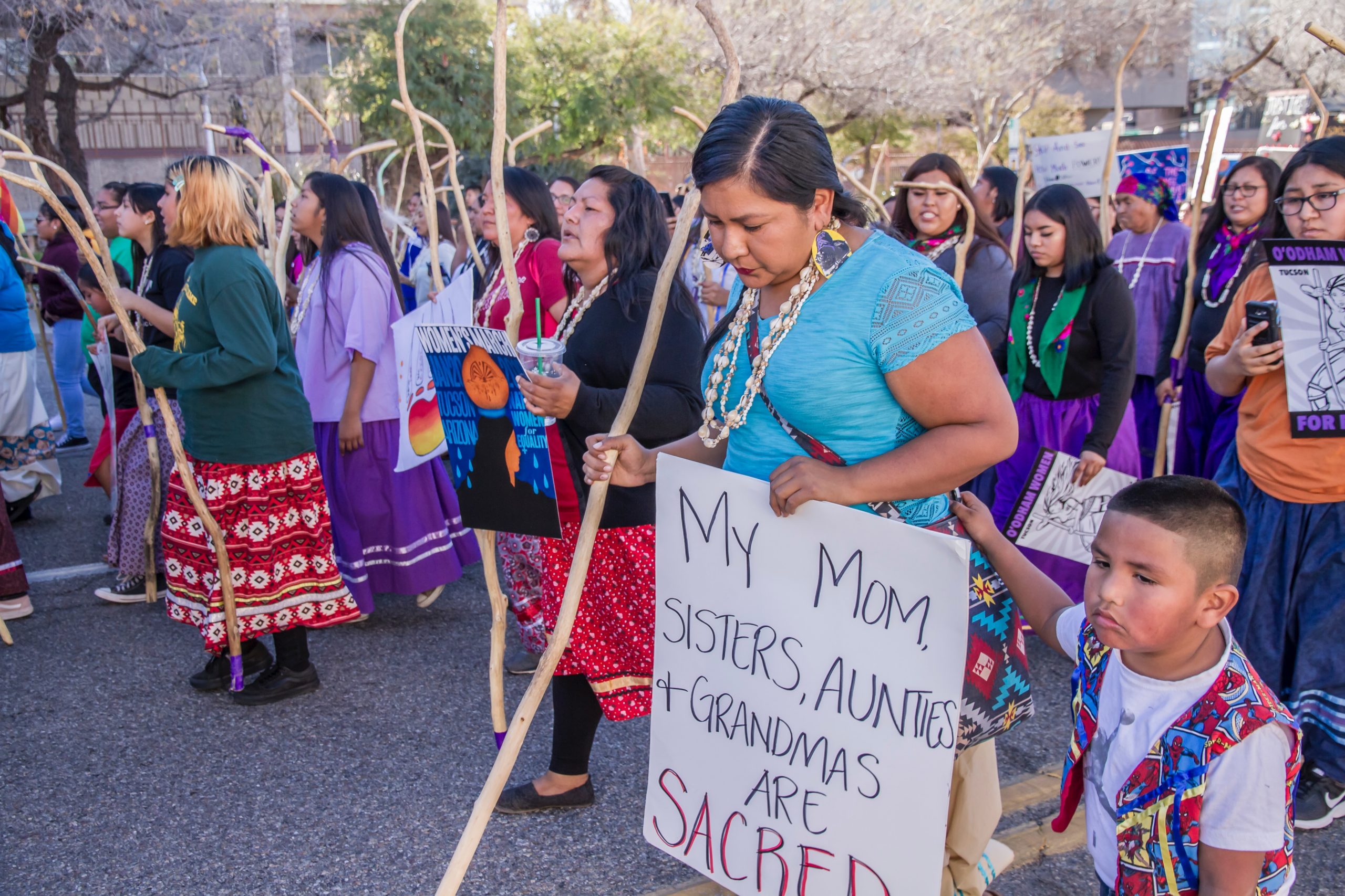 Indigenous Tohono Women led the Tucson 2019 Women’s March with a show of strength, resilience and power. This woman’s sign said: My Mom, Sisters, Aunties and Grandmas are sacred. Her son was by her side. International Women’s Day