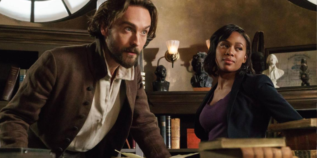 Tom Mison and Nicole Beharie as Ichabod Crane and Abbie Mills. They are in their library, leaning over books on the table. They are looking at someone from across the table. 